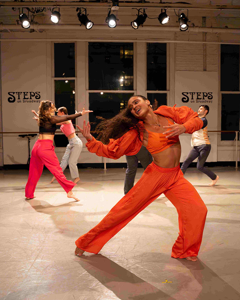 Dancers perform energetically in a studio with large windows. The foreground dancer wears a bright orange outfit, striking a dramatic pose. Three other dancers are in the background. The studio has STEPS on Broadway banners on the wall.