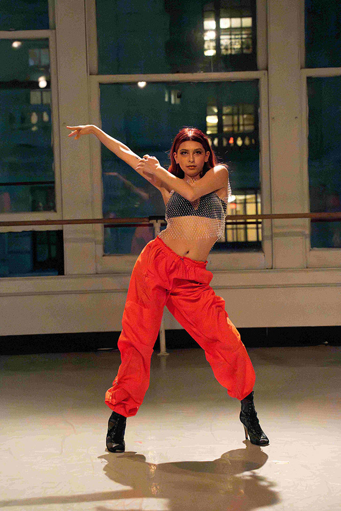 A person with red hair is dancing energetically in a studio. Theyre wearing a mesh top, red pants, and black boots. Large windows in the background show a glimpse of a city at night.