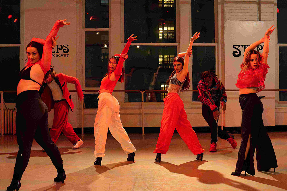 A group of dancers in vibrant outfits strike dynamic poses in a studio with large windows. They wear a mix of red, black, and white clothing, and the ambiance suggests energetic movement. The signs in the background read STEPS on Broadway.