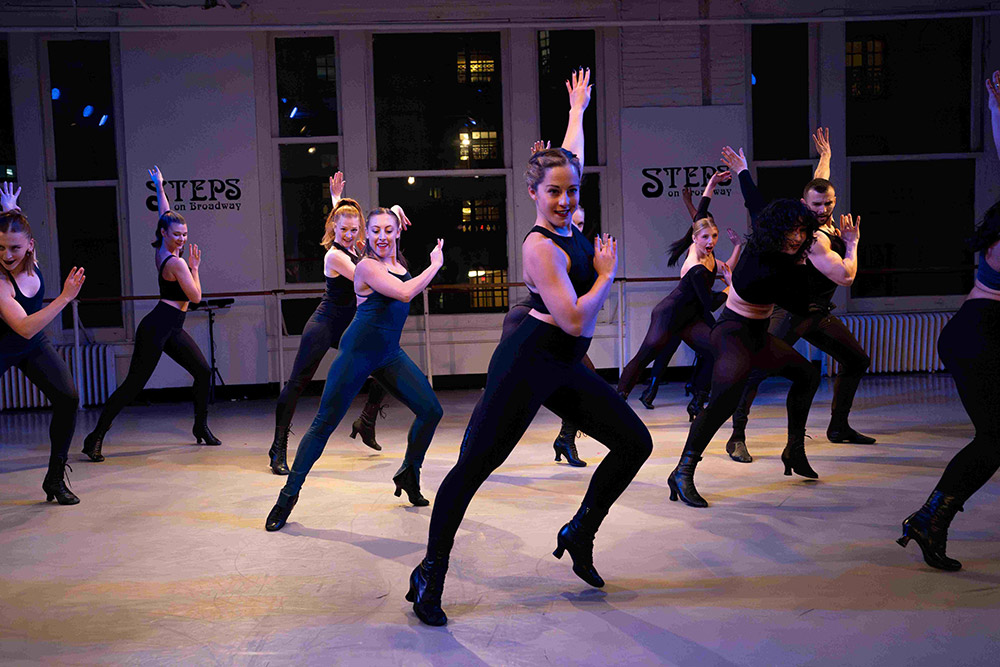 A group of dancers in form-fitting attire perform a synchronized routine in a studio with large windows. They are in dynamic poses, exuding energy and movement, with some signs reading Steps visible on the walls.