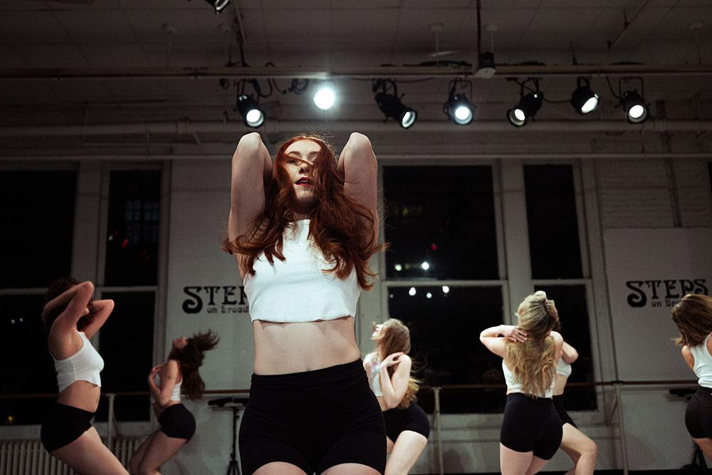 A group of dancers rehearses in a studio. The central dancer, wearing a white crop top and black shorts, poses confidently under bright overhead lights. Other dancers in similar outfits are blurred in the background, mimicking the movement.