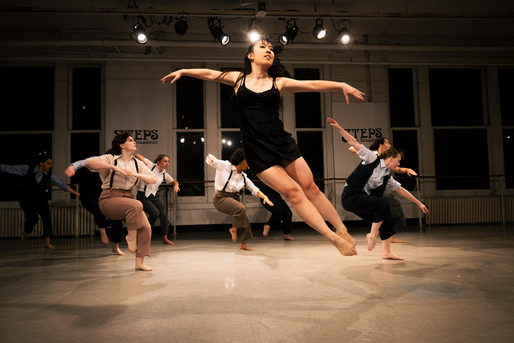 A group of dancers energetically performs on stage. The focus is on one dancer mid-air, arms extended. The room is dimly lit with spotlights, and others around are in motion, wearing casual dance attire.