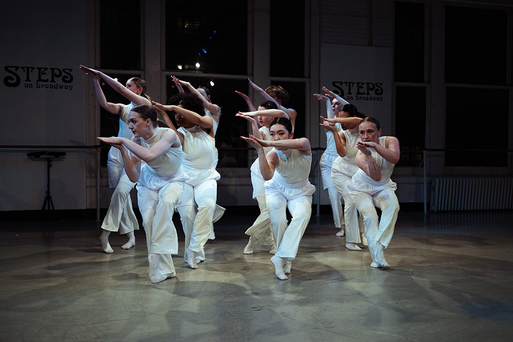 A group of dancers in white costumes perform a synchronized dance in a studio, with their arms extended and bent at the elbows, creating a dynamic V shape. The studio has large windows and Steps on Broadway is visible in the background.