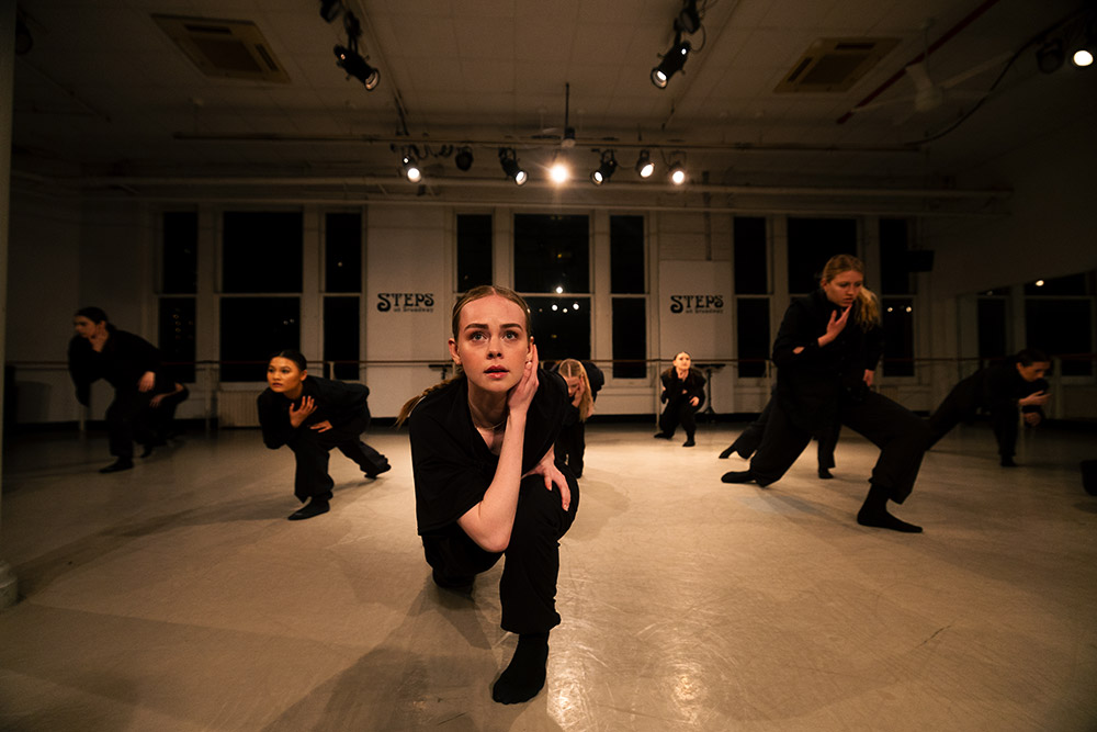 Dancers in black clothing perform a contemporary routine in a dimly lit studio. One dancer kneels in the foreground with others behind her striking dramatic poses against a backdrop of large windows and studio lights.