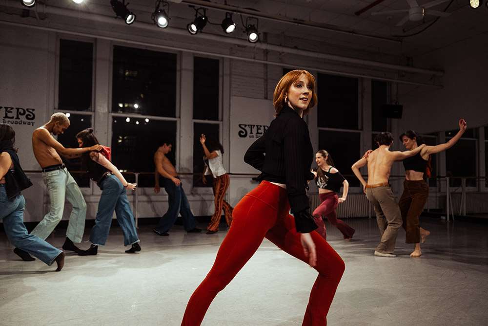 A group of dancers in a studio performing choreographed movements. They are wearing a variety of casual outfits, with one dancer in the foreground wearing red pants and a black top. Studio lights illuminate the space as they practice their routine.