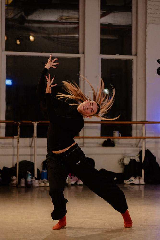 A person with long hair is dancing energetically in a studio, wearing a black outfit and red socks. Their hair is dramatically lifted as they move, with outstretched arms and a vivid expression. The background shows ballet barres and windows.