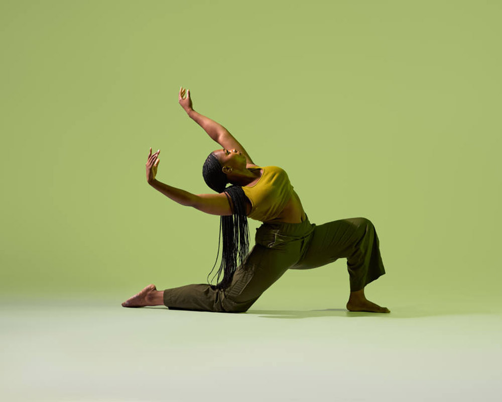 A person with long hair performs a graceful dance pose, lunging with one knee bent and the other leg extended. They wear a yellow top and green pants. The background is a solid light green.