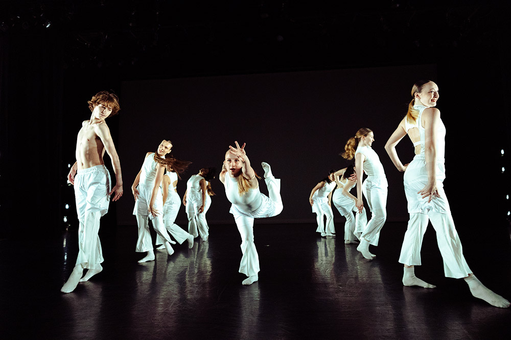 A group of dancers in white outfits performs on stage. One person is in the center, balancing on one leg with hands forming peace signs. The background is dark, highlighting the dancers movements.