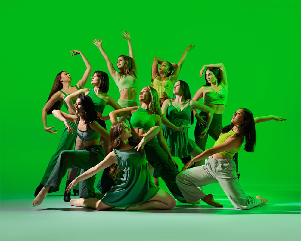 A group of dancers pose in artistic formation against a vibrant green background. They wear various shades of green clothing, showcasing a sense of movement and expression through their dynamic poses.