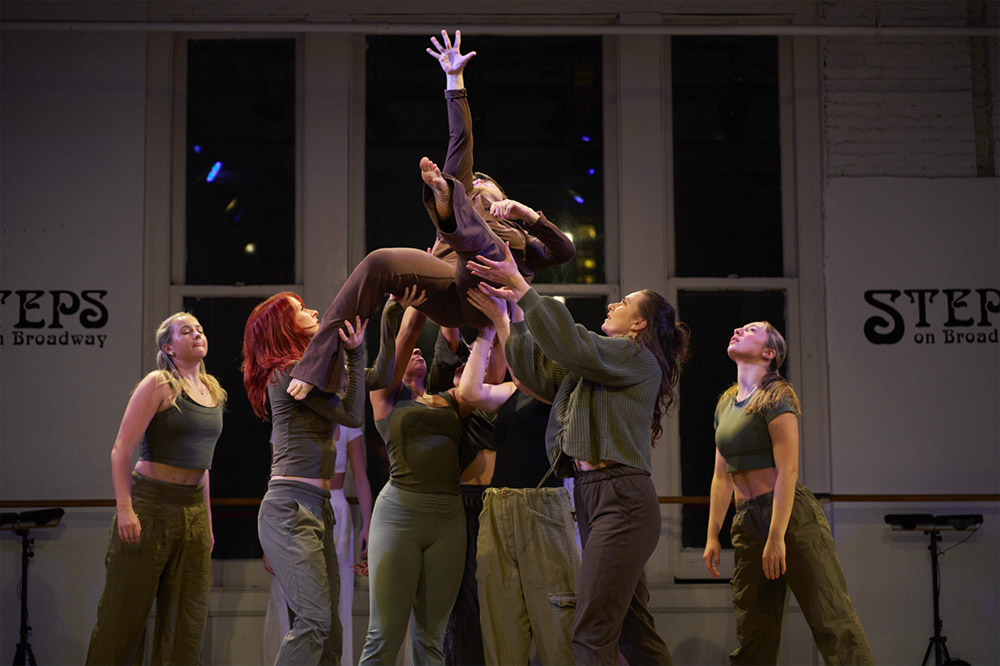 A group of dancers, dressed in earthy tones, lift a fellow performer high above their heads in a studio marked STEPS on Broadway. The scene captures a dynamic and graceful dance moment, with expressions of focus and collaboration.