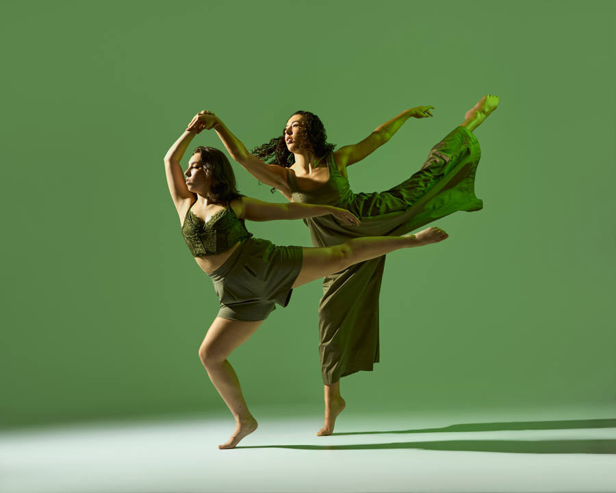 Two women dance gracefully against a green background. One balances on one leg with an arm extended, while the other leaps with an outstretched leg. Both wear earthy-toned clothing.