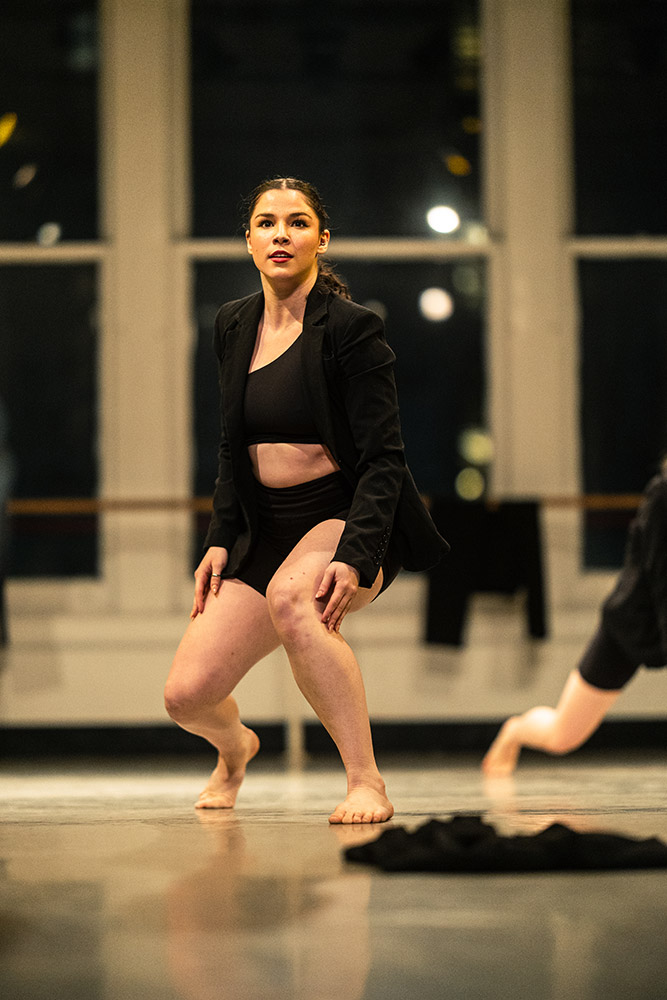 A dancer in a black outfit performs a movement in a brightly lit dance studio. She is barefoot and appears focused, with large windows in the background. The atmosphere conveys concentration and grace.