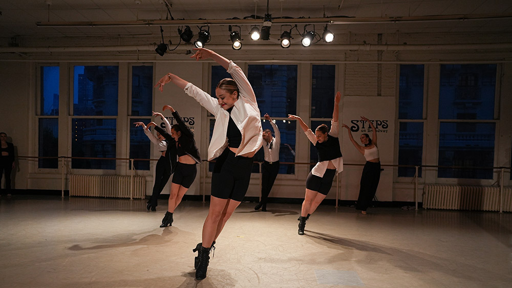 Dancers in a studio perform a choreographed routine with dynamic movements, wearing black and white attire. The room has large windows, and the stage is lit with overhead spotlights.