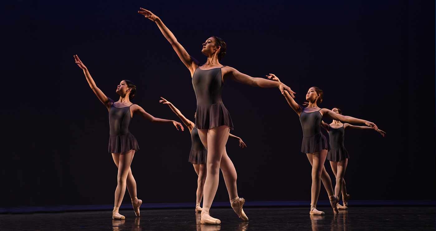 Five Pre-Professional Division dancers on a dark stage, arms raised, in brown dance dance skirts